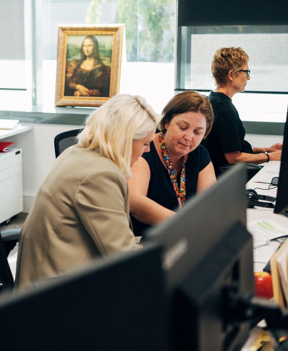 A bright office with two people working collaboratively, another person at their workstation in the background with the Mona Lisa leaning against the window.